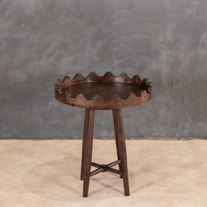 May include: A dark brown, round side table with a scalloped edge. The table has three legs and an X-shaped support near the base. The table is made of wood and has a natural finish.
