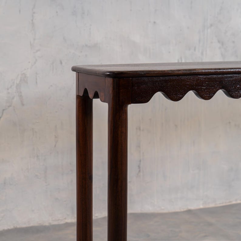 May include: A dark brown wooden table with a scalloped edge and four cylindrical legs. The table has a smooth, polished surface and stands against a textured, light gray wall.