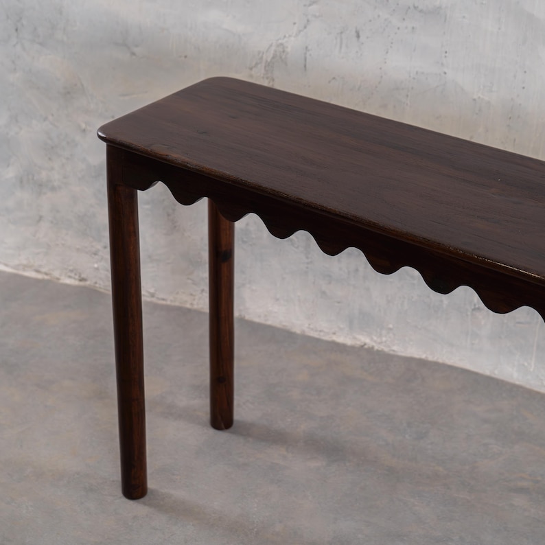 May include: A dark brown, narrow console table with a scalloped edge and four cylindrical legs. The table is made of wood and has a rectangular top with rounded corners. The legs are evenly spaced and provide support for the table.