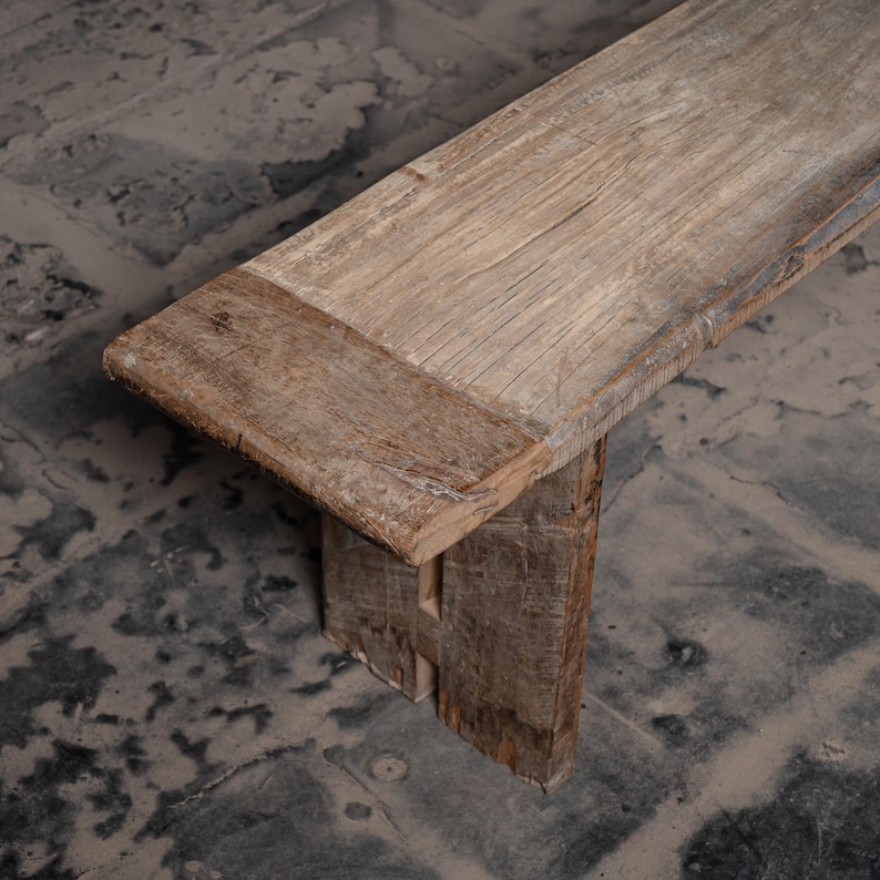 May include: A rustic wooden bench with a natural finish. The bench has a simple design with a wide, flat top and two sturdy legs. The wood has a weathered look, with knots and grain patterns visible.