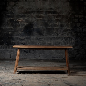 Peut inclure: Une console en bois rustique avec une seule étagère. La table a une finition brun foncé et est faite de bois de récupération. Elle est posée sur un sol en pierre grise rugueux.