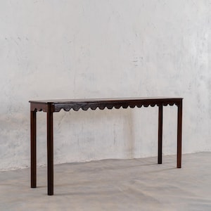 May include: A dark brown, narrow console table with a scalloped edge and four straight legs. The table is made of wood and has a simple, elegant design. The table is approximately 60 inches long.