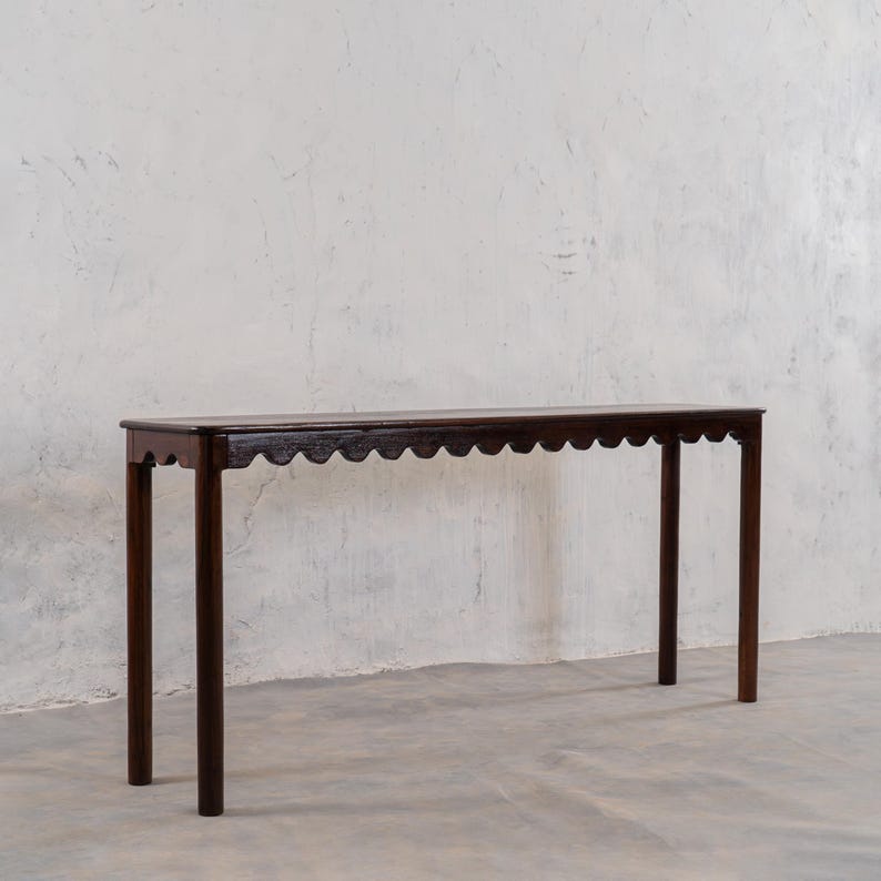 May include: A dark brown, narrow console table with a scalloped edge and four straight legs. The table is made of wood and has a simple, elegant design. The table is approximately 60 inches long.