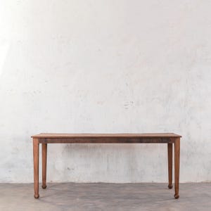 May include: A rustic wooden table with a natural brown finish and turned legs. The table is standing on a light gray concrete floor in front of a white wall.
