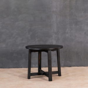 Op de afbeelding: Een kleine, ronde, zwarte houten bijzettafel. De tafel heeft een donkere afwerking en een eenvoudig, stevig ontwerp. Het tafelblad heeft een diameter van ongeveer 46 cm, ondersteund door vier poten die zijn verbonden door een kruisvormige beugel voor stabiliteit.