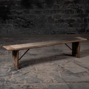 May include: A long, rustic wooden bench with a simple design. The bench has a natural wood finish and is supported by two sturdy legs with a metal brace.
