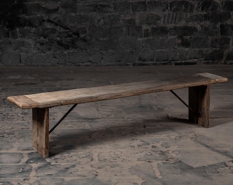 May include: A long, rustic wooden bench with a simple design. The bench has a natural wood finish and is supported by two sturdy legs with a metal brace.