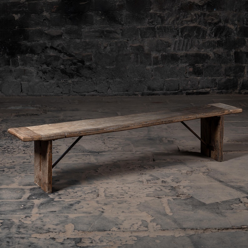 May include: A long, rustic wooden bench with a simple design. The bench has a natural wood finish and is supported by two sturdy legs with a metal brace.