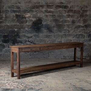 Puede incluir: Una consola larga de madera marrón oscuro con cuatro cajones y un estante inferior. La mesa tiene un estilo rústico y de granja.