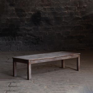 May include: A rustic wooden bench with a weathered grey finish. The bench has a rectangular top and four legs. It is sitting on a concrete floor in front of a dark stone wall.
