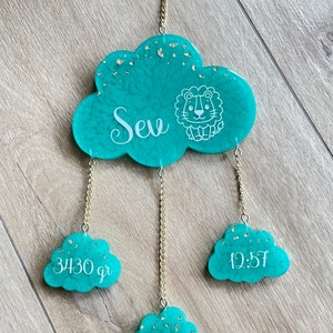 Op de afbeelding: Een turquoise wolkenvormige baby mobile met gouden accenten en een leeuwenillustratie. De mobile toont de naam "Seu", gewicht 3430 gram, lengte 50 cm en tijd 12:57. Het hangt aan een gouden ketting en ring.