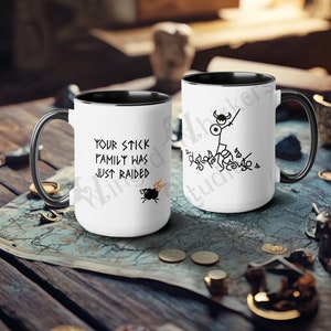 Puede incluir: Dos tazas de cerámica blanca con asas y bordes negros. Una taza tiene el texto "YOUR STICK FAMILY WAS JUST RAIDED" y un caldero de dibujos animados. La otra taza presenta un guerrero de palitos. Ambas tazas están sobre un mapa.