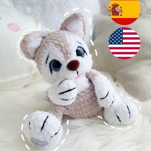 Puede incluir: Un perro de peluche tejido a crochet con pelaje blanco y marrón claro, ojos azules y nariz marrón. El perro está sentado sobre una superficie blanca y esponjosa. La imagen también incluye las banderas de España y Estados Unidos.