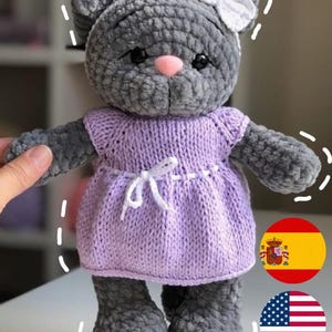 Puede incluir: Un juguete de gato de crochet gris que lleva un vestido morado con un lazo blanco. El juguete tiene una nariz rosa y ojos negros.