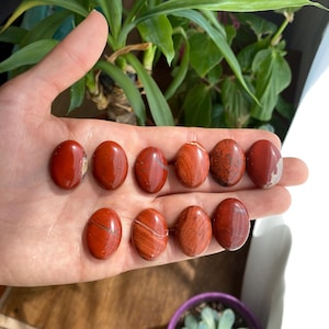 Peut inclure: Dix pierres de jaspe rouge roulées, chacune avec un motif unique de rayures rouges et blanches. Les pierres sont lisses et polies, et elles sont disposées en deux rangées sur une main.