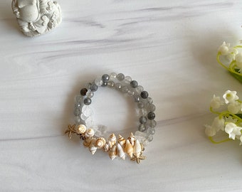 May include: Two beaded bracelets. One bracelet features gray and clear beads, while the other is adorned with small seashells and gold starfish charms. The bracelets are displayed on a white marble surface.