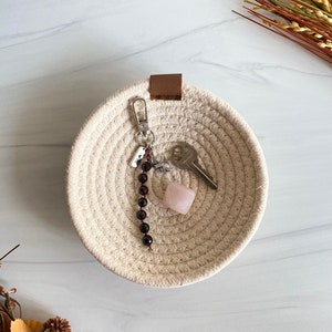 May include: A small, round, beige woven basket with a brown leather handle. The basket contains a pink crystal, a silver key, and a dark red beaded necklace.