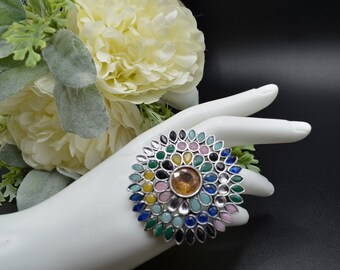 May include: A silver ring with a large, colorful stone in the center. The stone is surrounded by smaller stones in various colors, including green, blue, pink, and yellow.