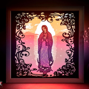 Könnte beinhalten: Gerahmtes Papierkunstwerk mit einer religiösen Figur in einem Gewand vor einem Sonnenuntergang. Das Kunstwerk ist von schwarzen, ausgeschnittenen Blumen- und Rankenmustern umrahmt, wodurch ein Schattenbox-Effekt entsteht.