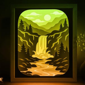 May include: A framed paper art piece depicting a waterfall scene. The artwork features layers of paper in shades of green, yellow, and brown, creating a 3D effect. The scene includes a waterfall, trees, mountains, and a sun in the sky.