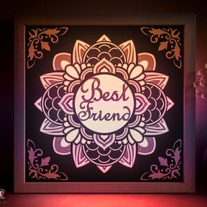 May include: A framed, layered paper art piece with a mandala design. The artwork features a central circle with the words "Best Friend" in a script font, surrounded by intricate floral patterns in shades of purple, brown, and cream. The frame is a light brown color.