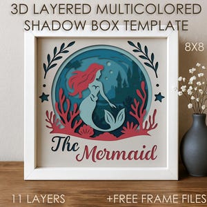 Könnte beinhalten: Gerahmte 3D-Schattenbox-Vorlage mit einem Meerjungfrau-Design. Das Kunstwerk zeigt eine Meerjungfrau mit roten Haaren und blauem Schwanz, umgeben von Korallen und Wasserelementen. Der Text "The Mermaid" ist in dekorativer Schrift dargestellt. Der Rahmen ist 20x20 cm groß.