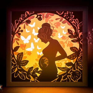 May include: A framed paper cut light box featuring a silhouette of a pregnant person, with an image of a fetus visible. The design includes butterflies and floral elements in shades of orange, yellow, and brown. The frame is made of wood.