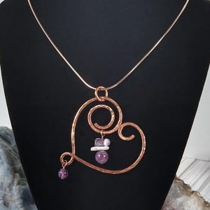 May include: A copper-colored heart pendant necklace. The heart-shaped pendant has a hammered texture and a spiral design. It is adorned with purple and white gemstones. The necklace is displayed on a black mannequin.