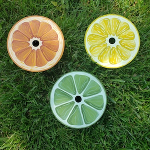 Puede incluir: Tres posavasos de cerámica pintados, cada uno representando una fruta cítrica diferente: naranja, limón y lima. Los posavasos son redondos y tienen un agujero en el centro.