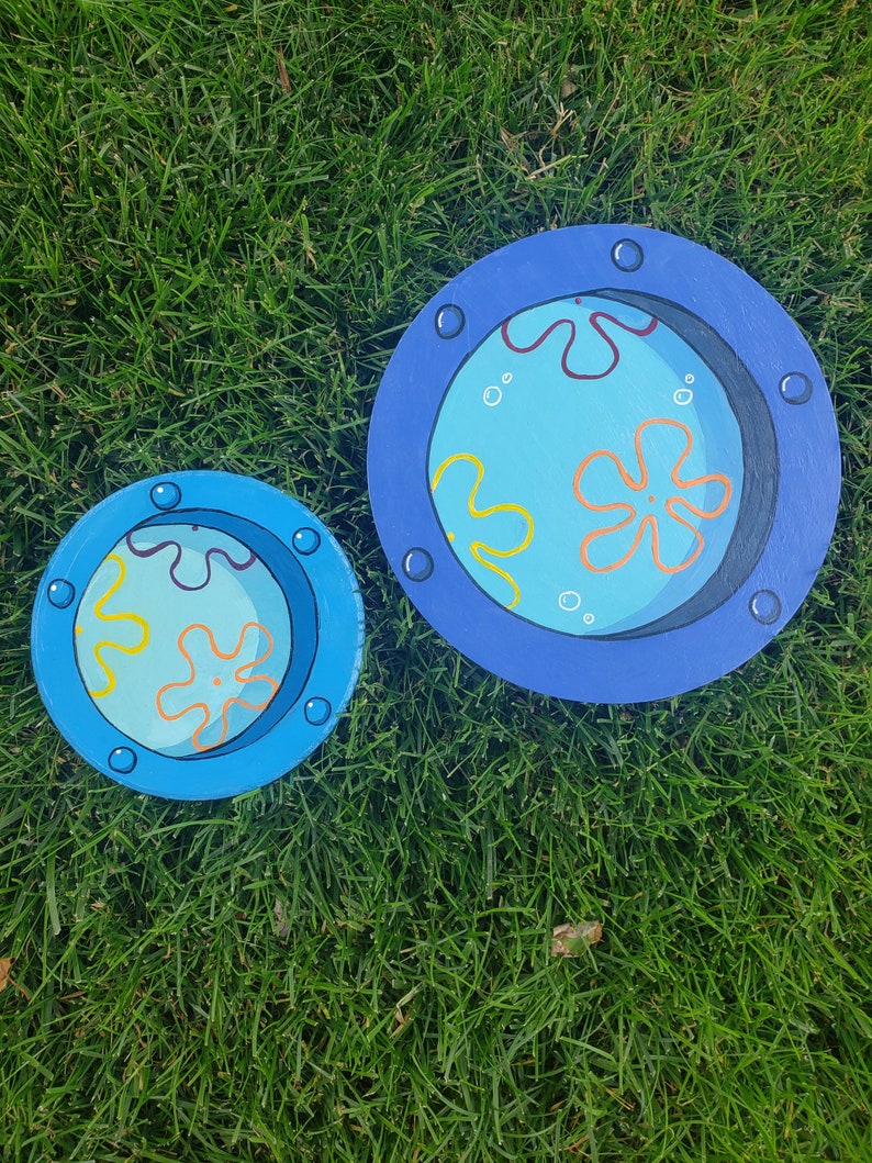 May include: Two round wooden circles painted to look like porthole windows. The larger circle is blue with a dark blue rim and has a yellow and orange starfish and yellow squiggly lines on a light blue background. The smaller circle is the same design but smaller.