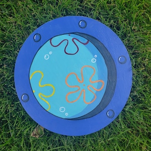 May include: A blue circular wooden sign with a painted image of a blue, yellow, and orange cartoon character with a black outline. The character is surrounded by white bubbles and is set against a black background. The sign has a blue border with white dots.