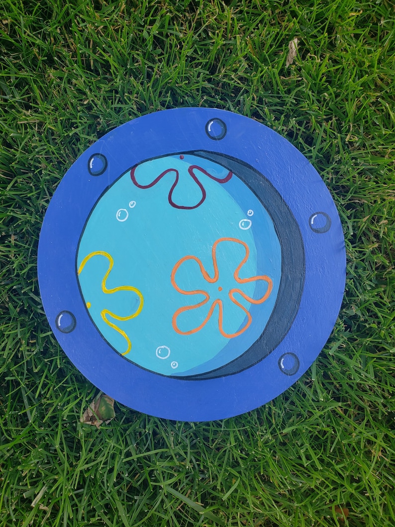 May include: A blue circular wooden sign with a painted image of a blue, yellow, and orange cartoon character with a black outline. The character is surrounded by white bubbles and is set against a black background. The sign has a blue border with white dots.