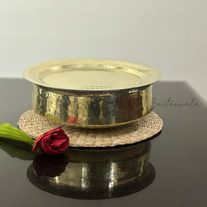 May include: A round, hammered brass bowl with a matching lid, featuring a reflective metallic gold finish and a textured surface. The bowl is placed on a circular woven mat, accompanied by a single red flower with a green stem.