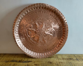 May include: A round copper tray featuring an intricate embossed floral pattern in the center, surrounded by a textured border with repeating geometric shapes and a scalloped edge. The metal surface has a polished, warm metallic finish throughout.