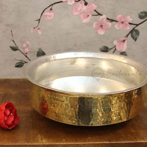 May include: A round, shallow metal bowl featuring a polished gold-tone exterior with a hammered texture and a smooth, silver-tone interior. The bowl has a wide, flat rim and displays the text Puri Bartanwala printed on the inner side.