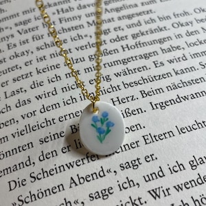 Könnte beinhalten: Zarte goldfarbene Halskette mit einem kleinen, runden Anhänger. Der Anhänger ist weiß mit einem handgemalten Design aus blauen Blumen. Die Halskette wird auf einer Buchseite präsentiert.