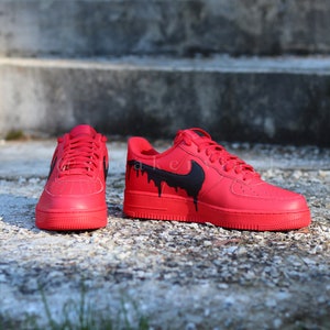 May include: Red sneakers with black accents. The shoes feature a solid red base with a black Nike swoosh and black dripping paint design. The sneakers are displayed on a textured surface.