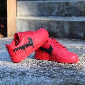 May include: A pair of red sneakers with black dripping paint design. The shoes have a classic low-top design, red laces, and a black Nike swoosh logo. The shoes are displayed on a textured surface.