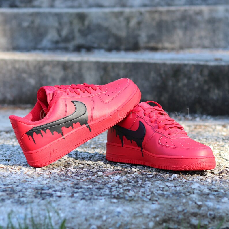 May include: A pair of red sneakers with black dripping paint accents. The sneakers have a black swoosh logo on the side and are displayed on a textured surface. The laces and soles are also red.
