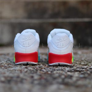 May include: A pair of white Nike Air Max 90 trainers with a red and green accent on the sole. The trainers have the Nike Air logo on the back.