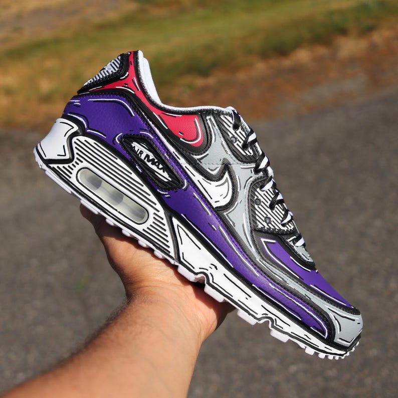 Puede incluir: Una zapatilla Air Max 90 morada, gris y roja con un dise&ntilde;o &uacute;nico delineado. La zapatilla presenta el logotipo "Air Max" y una suela blanca. El dise&ntilde;o incluye contornos negros, d&aacute;ndole una apariencia de dibujos animados. La zapatilla se sostiene en una mano.