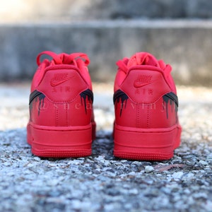 May include: Red Nike Air Force 1 sneakers with black drip design accents. The Nike logo is embossed on the heel. The sneakers are displayed on a textured surface.