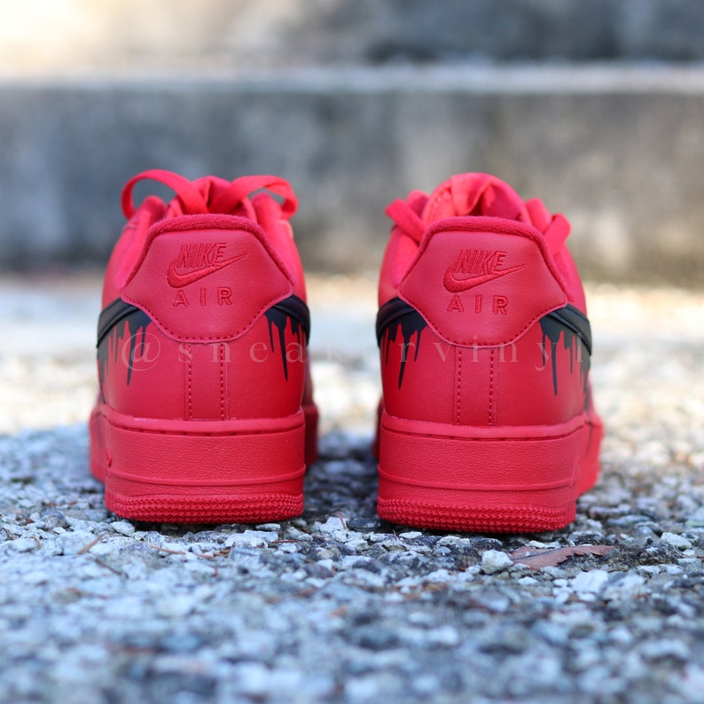 May include: Red Nike Air Force 1 sneakers with black drip design accents. The Nike logo is embossed on the heel. The sneakers are displayed on a textured surface.