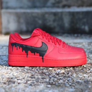 May include: Red Nike Air Force 1 sneakers with black dripping paint design. The iconic Nike swoosh is black. The word "AIR" is printed on the midsole. The laces and sole are red.