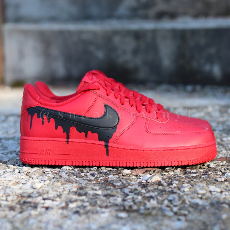May include: Red Nike Air Force 1 sneakers with black dripping paint design. The iconic Nike swoosh is black. The word "AIR" is printed on the midsole. The laces and sole are red.