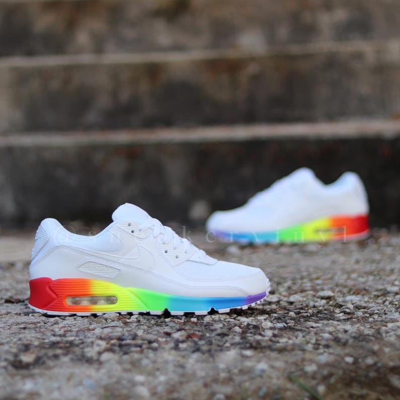 May include: White Nike Air Max 90 trainers with a rainbow coloured sole. The trainers are on a grey stone surface.