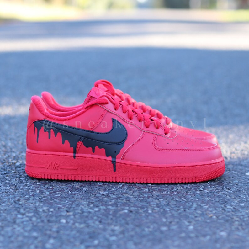 May include: Red sneakers with black dripping paint design. The sneakers have a black Nike swoosh and red laces. The word "AIR" is printed on the side of the sole. The shoes are on a gray surface.