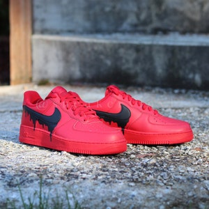 May include: A pair of red sneakers with black dripping paint design. The shoes have a black Nike swoosh and are displayed on a textured surface. The sneakers are a bold fashion statement.