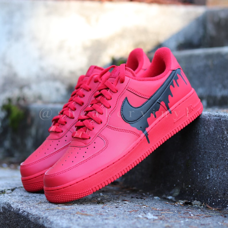 May include: A pair of red sneakers with black accents. The shoes feature a black Nike swoosh and black dripping paint design on the side. The word "AIR" is visible on the side of the sole. The sneakers are laced with red laces.