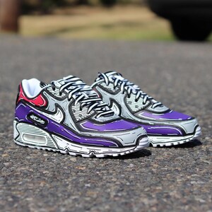 Puede incluir: Un par de zapatillas moradas, grises y blancas con contornos negros. Los zapatos tienen un dise&ntilde;o &uacute;nico con un acento rojo en el tal&oacute;n. Las zapatillas est&aacute;n sobre una superficie de asfalto.